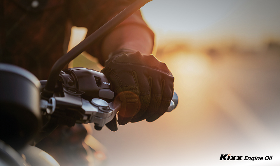 A rider’s gloved hand is held poised over the brakes of a motorcycle against a sunset background
