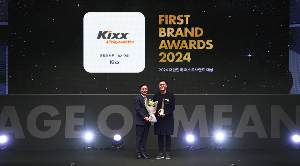 A host, left, onstage with a bouquet next to a GS Caltex representative holding a trophy, below signboards bearing the Kixx logo and the text “First Brand Awards 2024”