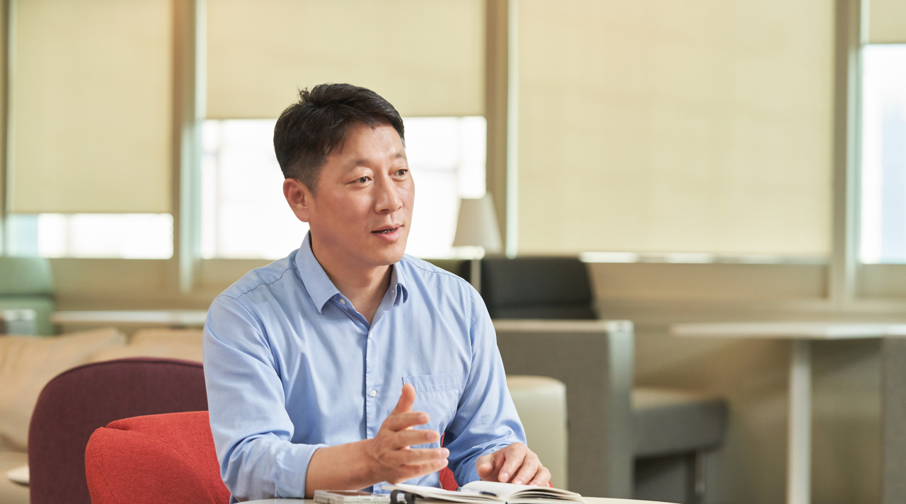 Kim Hyun Wook sits at a desk with a notebook and speaks to someone off-screen. 
