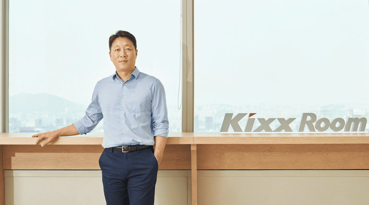 Kim Hyun Wook, dressed in business casual, looks into the camera as he stands next to a sign reading “Kixx Room.” 