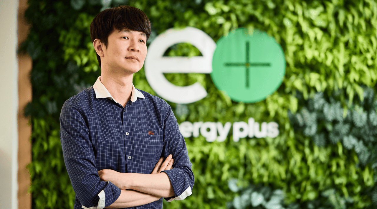 Kixx manager Kwan Hee Choi wears a blue button-up shirt and crosses his arms in front of a leafy wall with the sign “energy plus.”