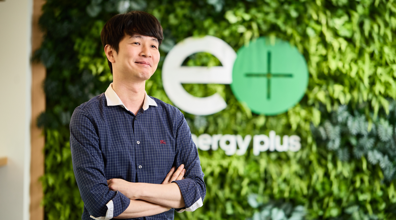 Kwan Hee Choi smiles as he crosses his arms in front of a leafy wall.