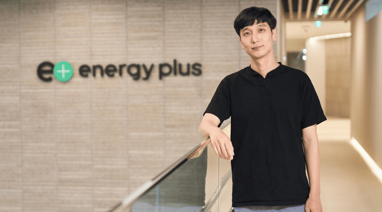 Rae Wook Cho, dressed casually, looks into the camera as he stands with his right arm resting on a railing, in front of “Energy Plus” signage.  