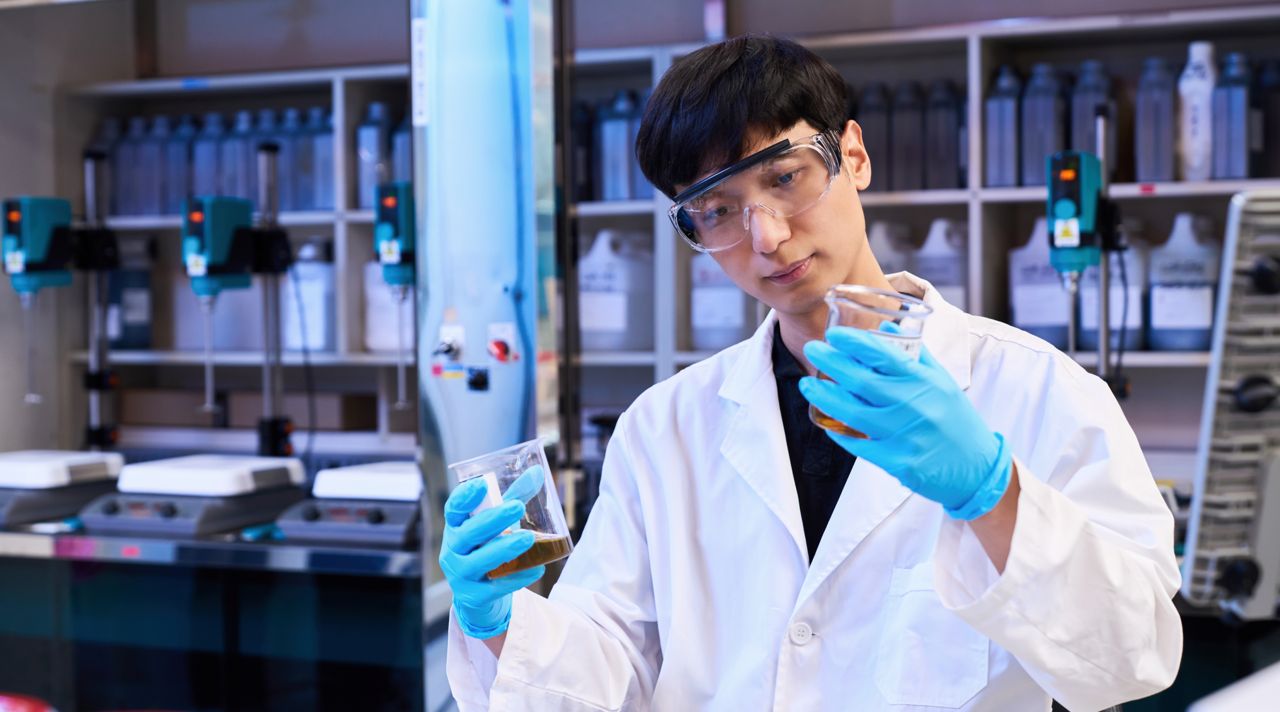 Rae Wook Cho in a laboratory, dressed in a white lab coat and wearing rubber gloves and safety goggles, examining substances in beakers.  