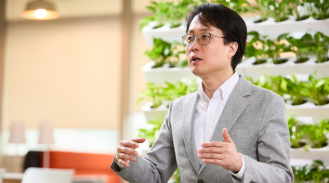 Changbeom Lee wears a grey suit and stands as he speaks animatedly, his arms raised in a gestural pose in a room filled with plants.