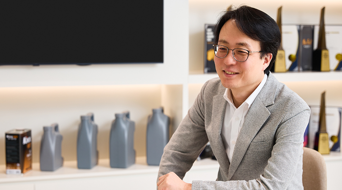 Changbeom Lee smiles and wears a grey suit, sitting with an arm placed on a small table in a room filled with Kixx products and awards.