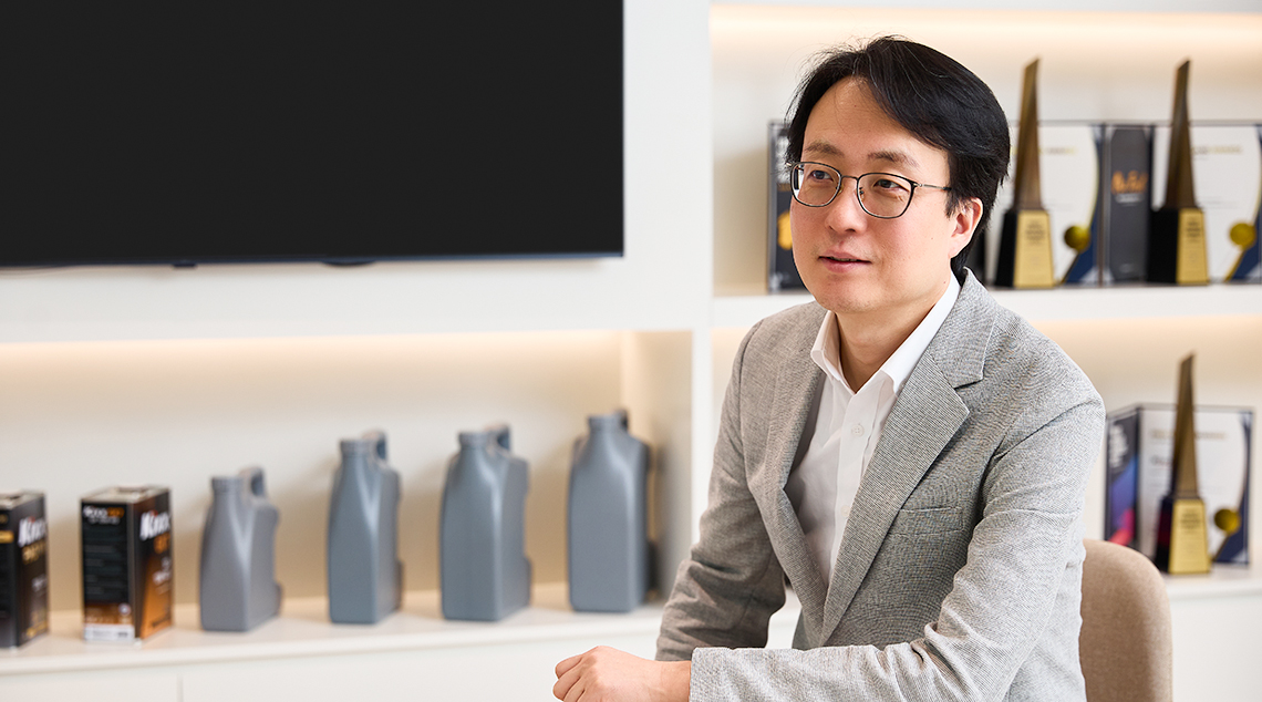 Changbeom Lee wears a grey suit and sits with an arm placed on a small table in a room filled with Kixx products and awards.