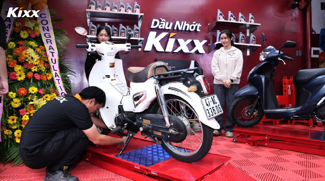 A Kixx technician works on a motorcycle at a Kixx Station while two female customers look on. 